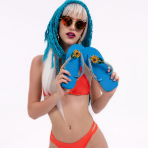 Chanclas Azules Flor Fotografía de estudio sobre fondo blanco con modelo de cabello rubio platino y bikini naranja neón de dos piezas. La modelo luce gafas de sol redondas de montura dorada y un pañuelo de crochet azul turquesa en la cabeza. Sostiene y exhibe un par de chanclas azul turquesa decoradas con girasoles de cristal amarillo y piedras preciosas verdes.